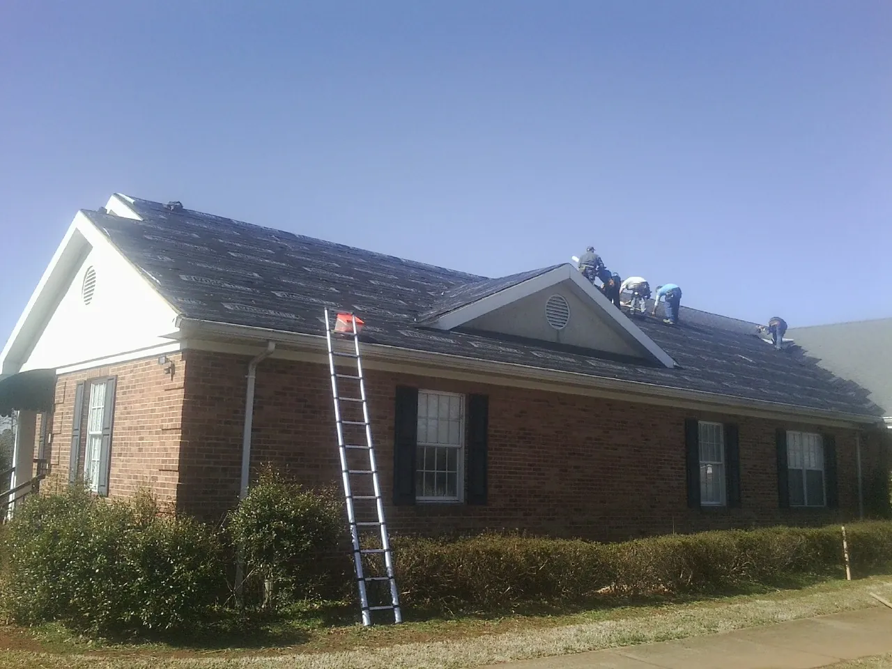 Crew tearing shingles off a brick ranch roof with a ladder set — Emergency Roof Repair work by our crew in Lanexa