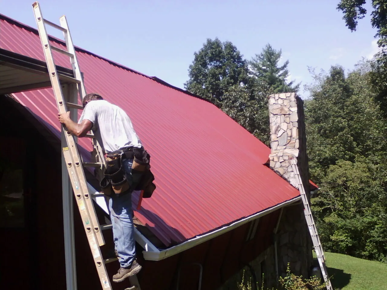 Roofer on a ladder beside a red metal roof and stone chimney — Soffit & Fascia Repair by Roofing Brothers in Lanexa